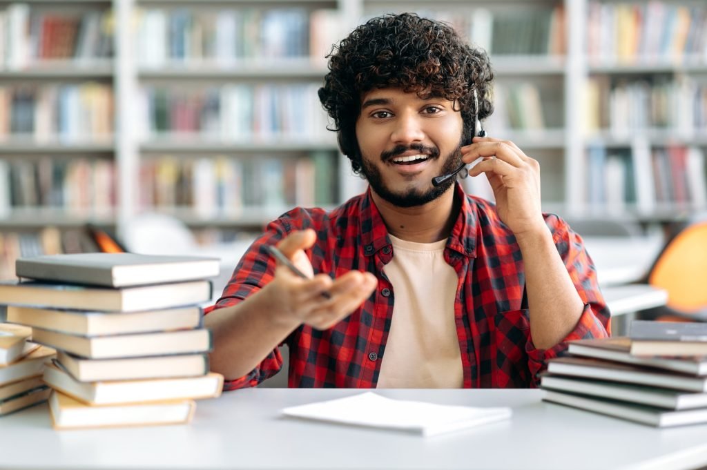 Positive Arabic or Indian university student with curly hair, wearing a headset, sits at a table in